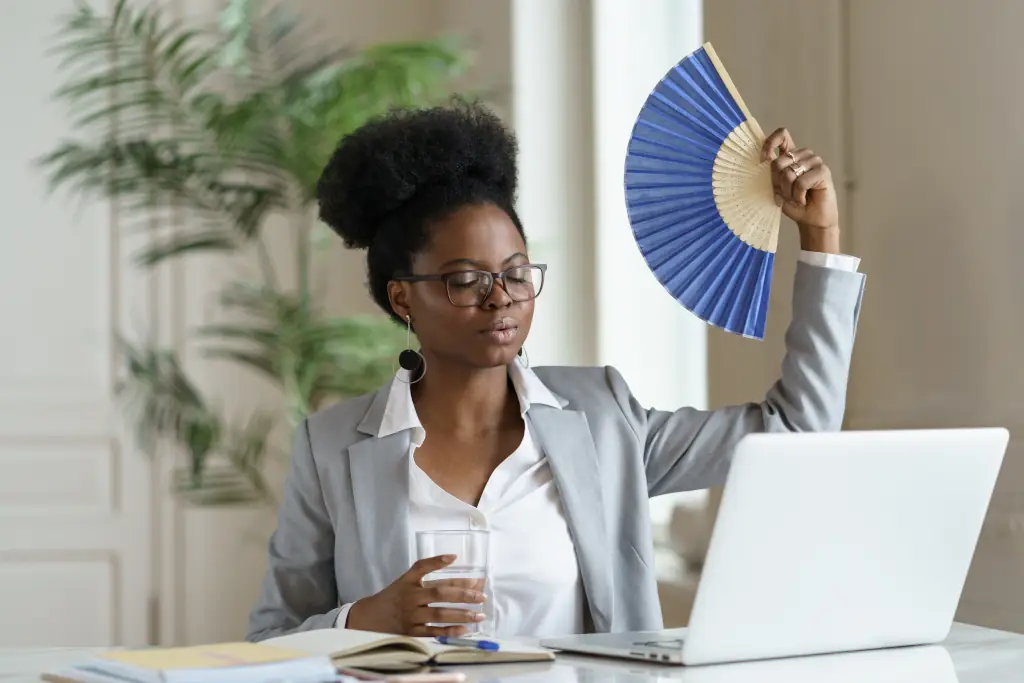 Erschöpfte Geschäftsfrau müde von Hitze winkendem Papierventilator für frische Luft am Arbeitsplatz Arbeit am Laptop